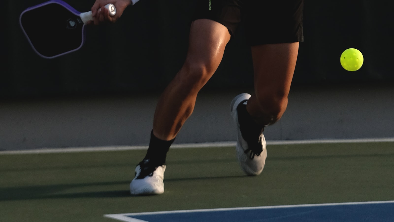 Close-up of a pickleball player's legs and paddle mid-swing, chasing down a ball on a dimly lit court